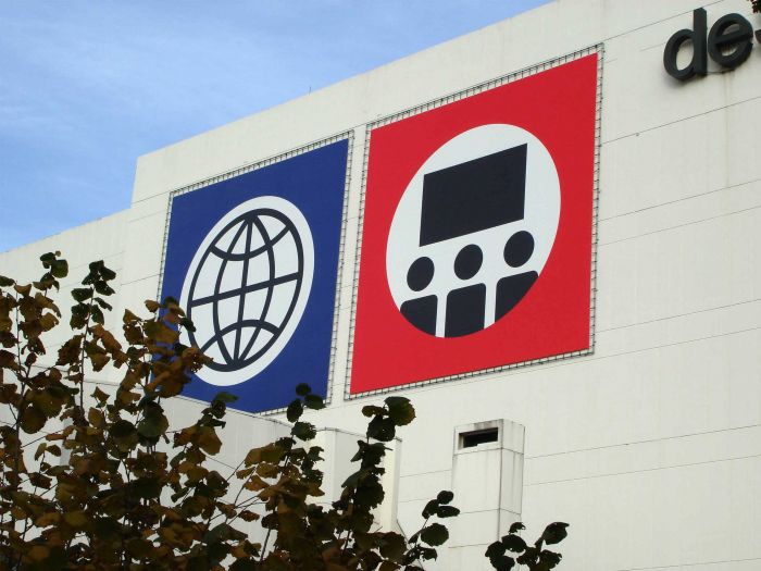 Zwei große quadratische Werbetafeln an der Wand eines Gebäudes. Die linke Tafel zeigt ein Symbol für das Internet (Globus), während die rechte Tafel ein Symbol für eine Präsentation oder ein Meeting (Bildschirm mit Personen) zeigt.