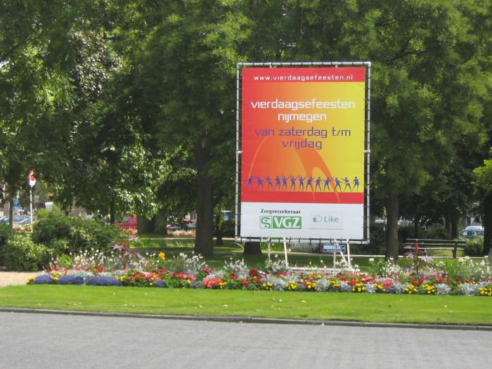 Ein großes, bunter Banner, das für die "Vierdaagsefeesten" (Vier-Tage-Fest) in Nijmegen werbt, das von Samstag bis Freitag dauert, mit buntem Design und Sponsorenlogos.