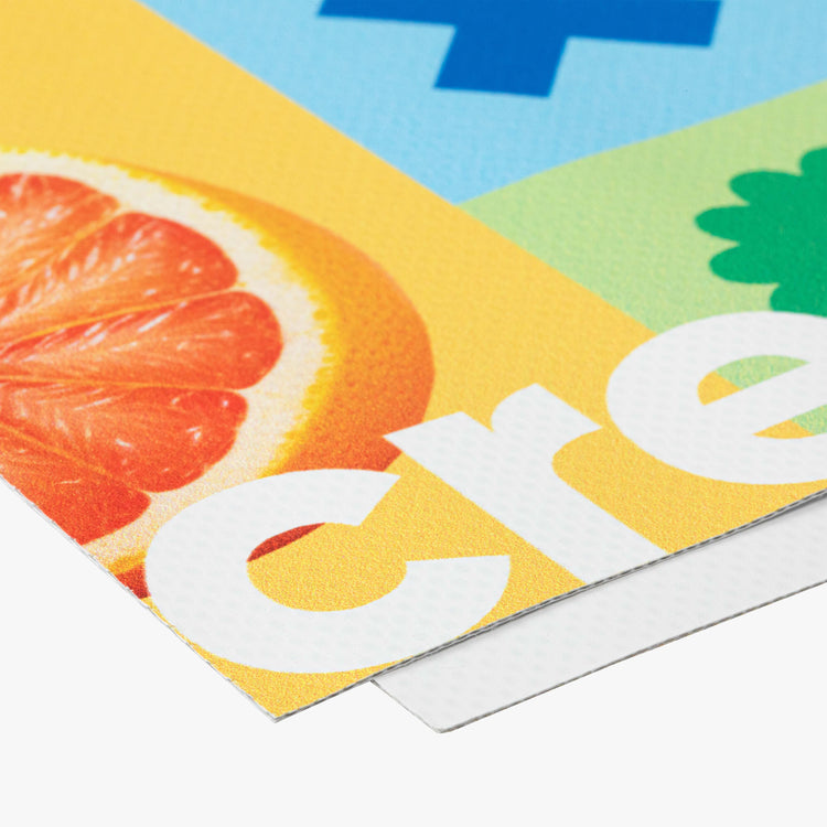 Dieses Bild zeigt einen detaillierten Ausschnitt eines bedruckten Materials, möglicherweise eines Banners oder Posters. Es ist zu erkennen, dass es sich um ein beschichtetes Material handelt, das mit einer fruchtigen Illustration (eine Grapefruit) und einem leuchtend gelben Hintergrund bedruckt ist. Die Textur des Druckmaterials ist gut sichtbar, mit einer fein strukturierten Oberfläche. Der Druck selbst scheint lebendige Farben zu verwenden.