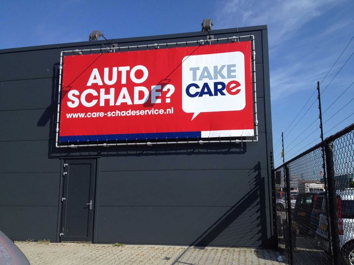 Ein großes Werbebanner an einem Gebäude, auf dem "AUTO SCHADE?" steht, mit dem Logo von TAKE CARE und einer URL für den Schadensservice. Das Banner hat einen roten Hintergrund mit weißer und blauer Schrift.