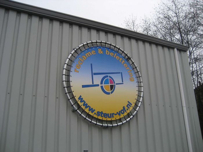 Ein rundes Werbeschild auf einer Gebäudewand, das den Text "reclame & belettering" und die URL "www.steur-vof.nl" zeigt. Das Schild hat einen blau-gelben Farbverlauf und ein grafisches Logo in der Mitte.