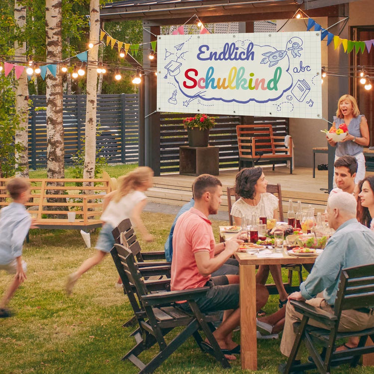 Einschulungs-Banner mit der Aufschrift „Endlich Schulkind“ im Garten aufgehängt, während eine Familie eine festliche Feier mit Essen und spielenden Kindern genießt.