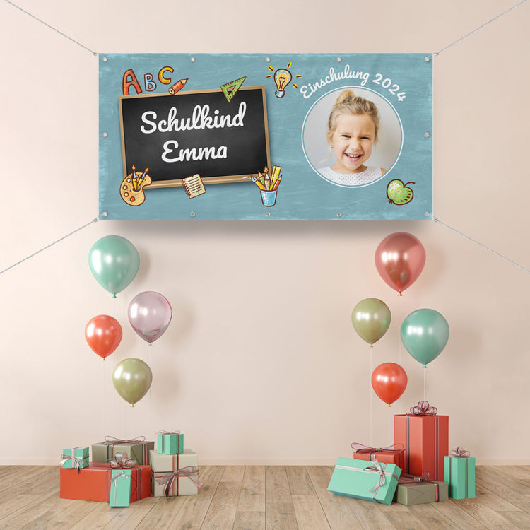 Ein selbst personalisierter Einschulungsbanner mit Ösen und Befestigung in einem geschmückten Raum. Der blaue Banner ist vorgestaltet und kann mit eigenem Namen und Foto gedruckt werden. Als Beispiel hier für das Schulkind Emma zur Einschulung 2024, mit bunten passenden Designs.