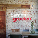 Das Bild zeigt einen Raum mit einer Backsteinwand, auf der die Worte 'Samen groeien' in weißer und roter Schrift an der Wand angebracht sind. Im Vordergrund stehen ein Tisch mit Stühlen und eine Vase mit Blumen.
