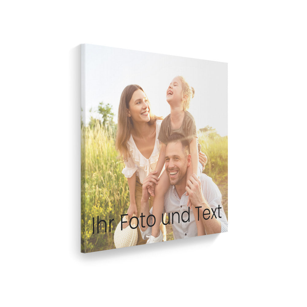 Ein Foto auf Leinwand, das eine glückliche Familie zeigt. Der Vater trägt das Kind auf den Schultern, während die Mutter an seiner Seite steht. Alle lächeln und genießen den Moment in der Natur. Unter dem Bild steht der Text "Ihr Foto und Text", was darauf hinweist, dass das Bild personalisiert werden kann. Die Szenerie ist von Sonnenlicht erleuchtet und im Hintergrund ist grünes Gras sichtbar.