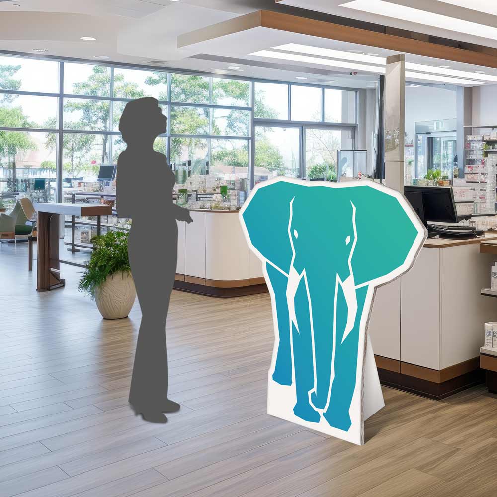 Der formgeschnittene Pappaufsteller in Form eines Elefanten steht für einen Größenvergleich neben einer Schattenfigur in Kassenbereich.