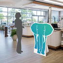 Der formgeschnittene Pappaufsteller in Form eines Elefanten steht für einen Größenvergleich neben einer Schattenfigur in Kassenbereich.