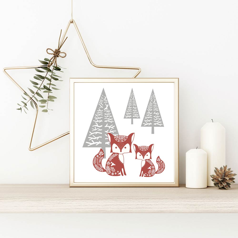 Ein eingerahmtes Bild mit zwei Füchsen und Tannenbäumen in Rot und Grau. Neben dem Bild stehen zwei weiße Kerzen und ein Tannenzapfen auf einem Regal, mit einem goldenen Stern und Zweigen im Hintergrund, passend zur Weihnachtsdeko.