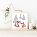 Ein eingerahmtes Bild mit zwei Füchsen und Tannenbäumen in Rot und Grau. Neben dem Bild stehen zwei weiße Kerzen und ein Tannenzapfen auf einem Regal, mit einem goldenen Stern und Zweigen im Hintergrund, passend zur Weihnachtsdeko.