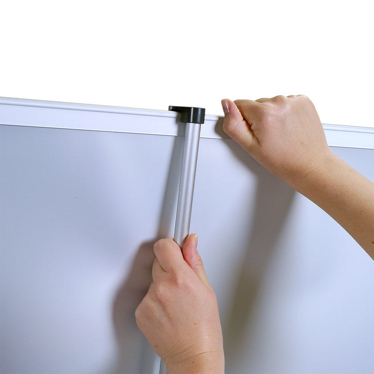 Eine Person fixiert einen Roll-Up-Banner, indem sie das Stützrohr an den oberen Rand des Banners anbringt. Die Hand der Person hält das Rohr fest, um es in die dafür vorgesehene Position zu bringen, um das Banner vollständig aufzubauen. Das Banner hat einen klaren weißen Hintergrund und wird mit einer stabilen, silbernen Metallstange montiert.