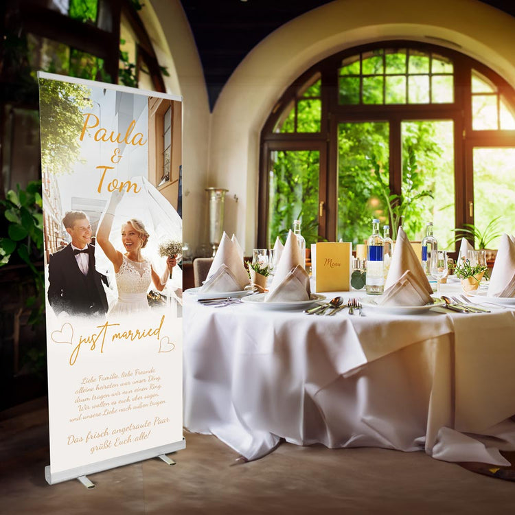 Ein Roll-up-Banner zur Hochzeit von Paula und Tom, das das frisch verheiratete Paar zeigt, wie sie fröhlich ihre Hände in die Luft werfen. Der Banner enthält den Text "just married" und eine persönliche Nachricht des Paares an ihre Familie und Freunde. Die Hochzeitstafel im Hintergrund zeigt ein elegantes, festlich gedecktes Esstischarrangement mit Menükarten, Gläsern und Pflanzen in einer stilvollen Location mit viel natürlichem Licht.