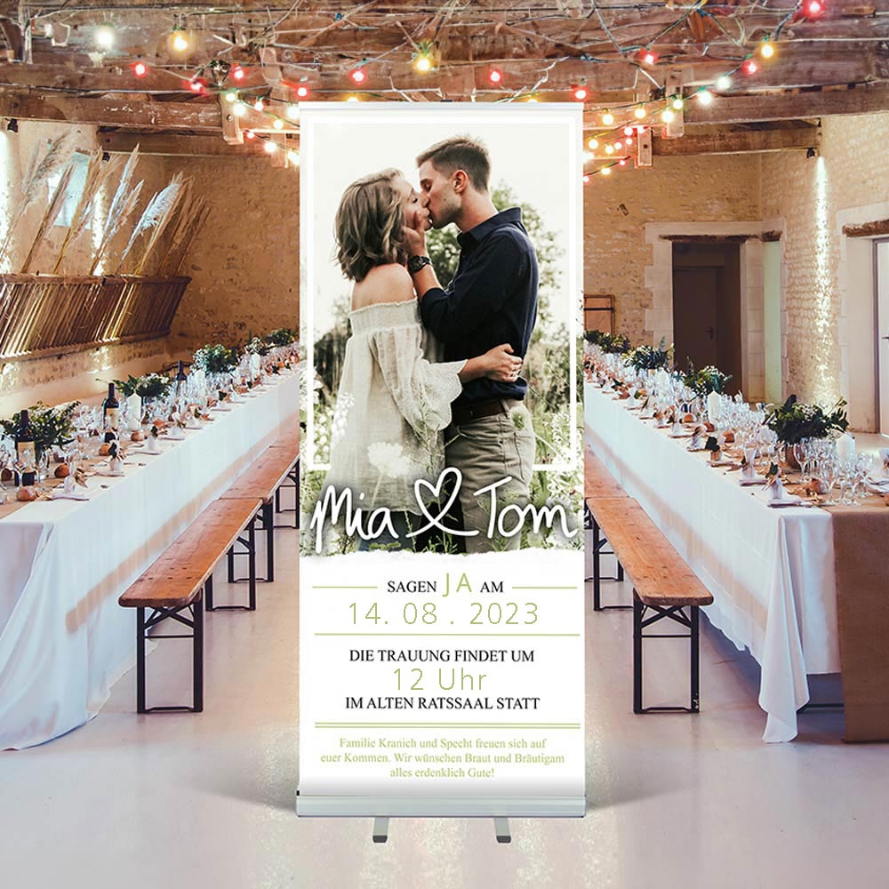 Ein stilvolles Roll-up-Banner mit einem romantischen Bild von einem Paar, das sich küssend in der Natur umarmt. Es ist eine Einladung zur Hochzeit von Mia und Tom. Das Banner enthält wichtige Details der Hochzeit, wie das Datum (14.08.2023), die Uhrzeit (12 Uhr) und den Veranstaltungsort (Alter Rathaussaal). Im Hintergrund sieht man eine festlich gedeckte Tafel in einer charmanten Veranstaltungslocation mit bunten Lichtern, die eine warme, einladende Atmosphäre schaffen.