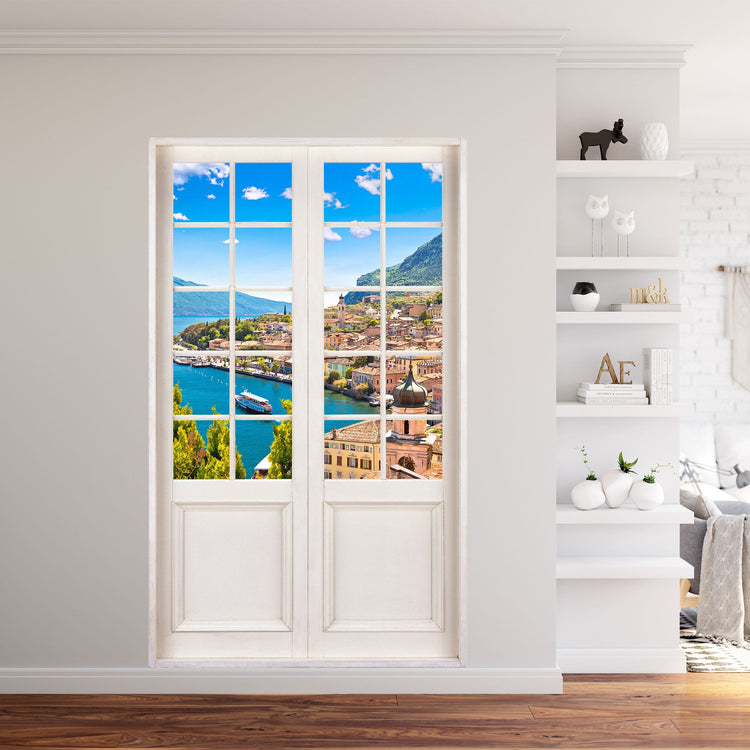 Selbstklebende Wandtapete Terassentür mit toller Aussicht auf ein Dorf am See. Verklebt in einem Wohnzimmer an der Wand neben einem Regal.