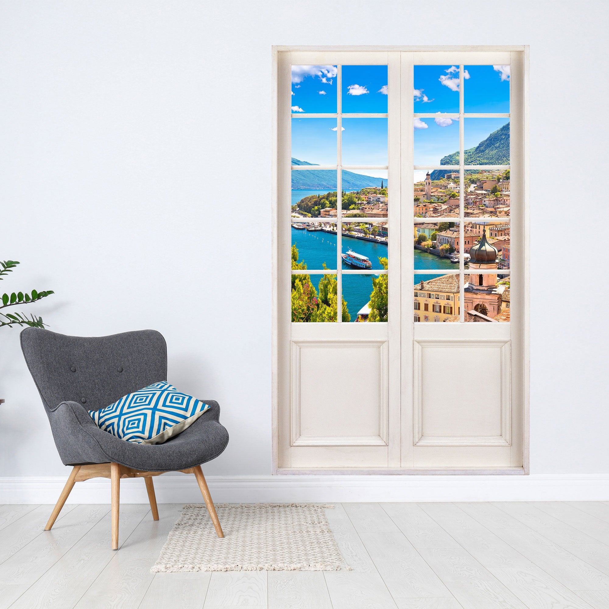 Gedrucktes Wandbild für Tapete und Möbel, mit toller Aussicht auf türkisfarbenen See und einem Dorf. Der Terrassentür Aufkleber ist an einer weissen Wand neben einem grauen Sessel verklebt.