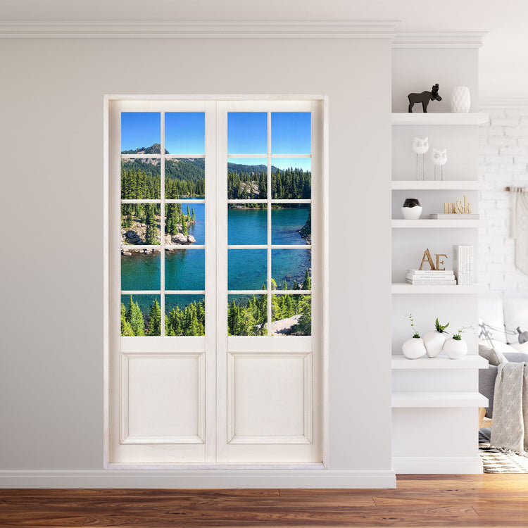 Selbstklebende Terrassentür mit Urlaubsmotiv, Tür mit weissem Sprossenfenster und Foto mit einem Bergsee, grünen Tannen und Bergen. Als Dekoidee in einem weissen Wohnzimmer neben einem Regal verklebt.