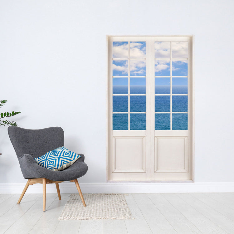 Wandaufkleber Terrassentür für Wohn- und Schlafzimmer, vergrößert Ihr Zimmer optisch. Hier mit dem Ausblick auf das Meer mit blauem Himmel, verklebt neben einem grauen Sessel als Dekoidee.