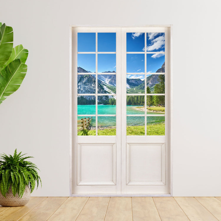 Wandaufkleber mit den Foto einer Terrassentür und Blick auf den italienischen Pragser Wildsee. An einer weissen Wand neben großen Zimmerpflanzen verklebt.