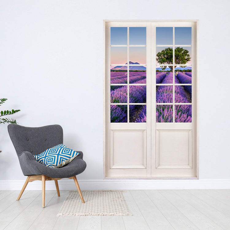 Selbstklebende Wandtapete mit einem Türrahmen und toller Aussicht auf ein Lavendelfeld. Als Dekoidee an einer weisen Wand verklebt, neben einem grauen Sessel.