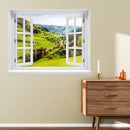 Offenes Fenster mit Ausblick auf grüne Hügel, ein idyllisches Dorf und bewaldete Berge – dekoratives Fensterbild mit realistischer 3D-Optik für naturnahes Wohnambiente. Als Dekoidee an einer gelben Wand neben einer Kommode aus Holz verklebt.