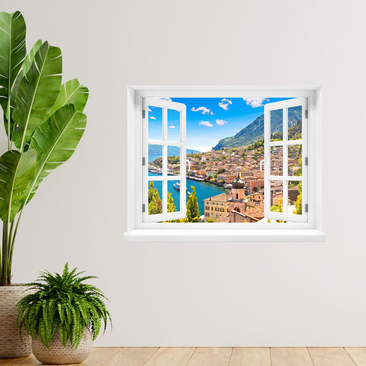 Wandsticker offenes Fenster "Dorf am See" auf heller Wand neben einer großen Zimmerpflanze.