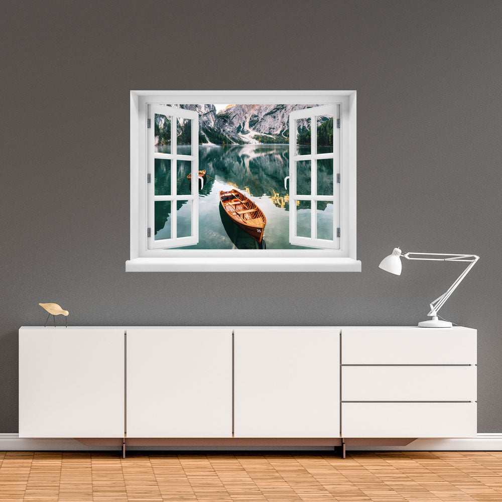 Wandaufkleber-Ansicht-offenes Fenster mit Blick auf ruhigen Bergsee mit Holzruderbooten, umgeben von steilen Felswänden und Wäldern – stimmungsvolle Naturkulisse in den Alpen. Als Dekoidee an einer grauen Wand, über einem weissen Sideboard verklebt.