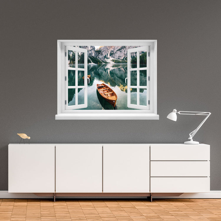 Wandaufkleber-Ansicht-offenes Fenster mit Blick auf ruhigen Bergsee mit Holzruderbooten, umgeben von steilen Felswänden und Wäldern – stimmungsvolle Naturkulisse in den Alpen. Als Dekoidee an einer grauen Wand, über einem weissen Sideboard verklebt.