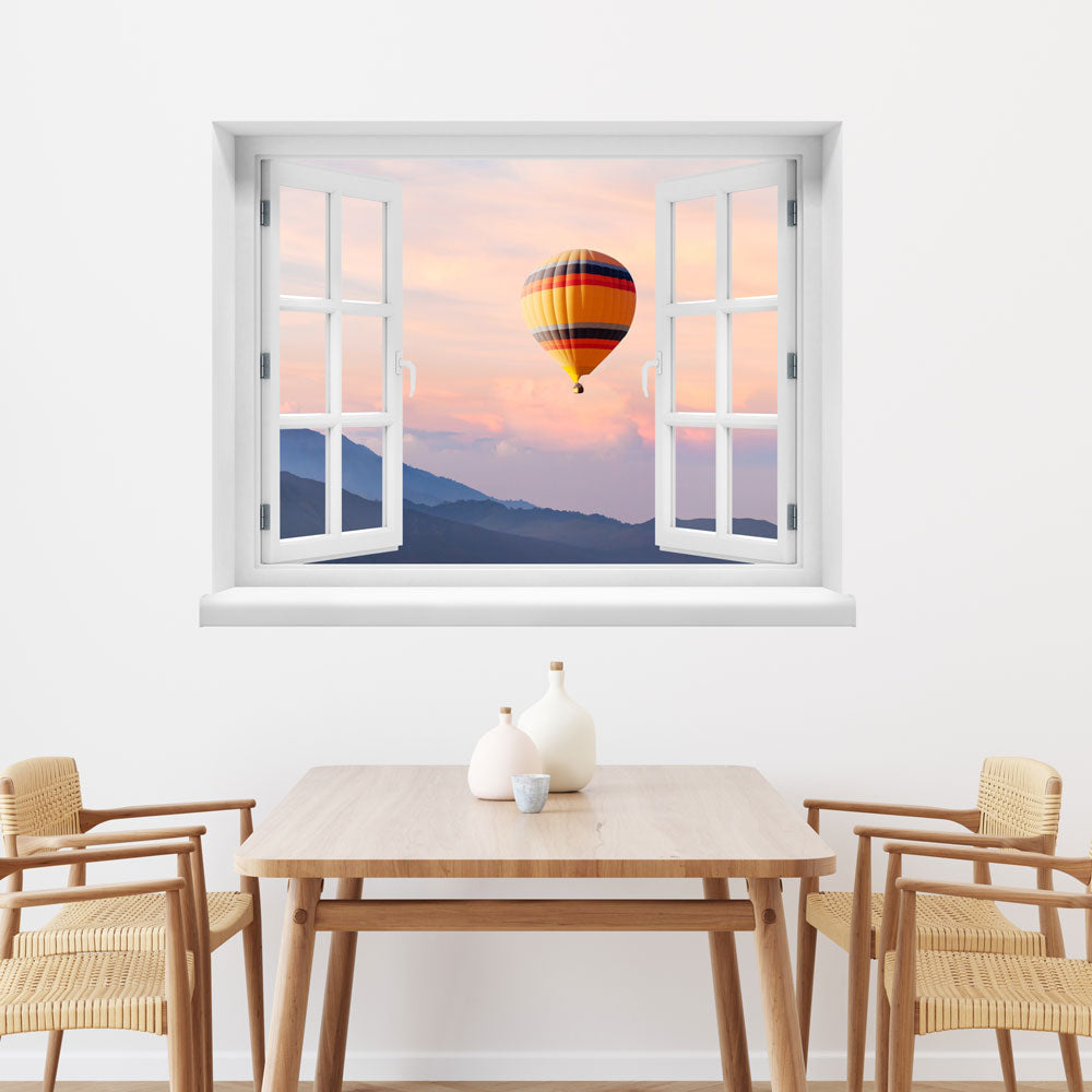 Wandaufkleber mit dem Motiv offenes Fenster mit Blick auf einen Heißluftballon im Sonnenuntergang über einer Berglandschaft – dekoratives 3D-Fensterbild mit Panorama-Effekt. Als Dekoidee über einem Esstisch aus Holz an einer weissen Wand.