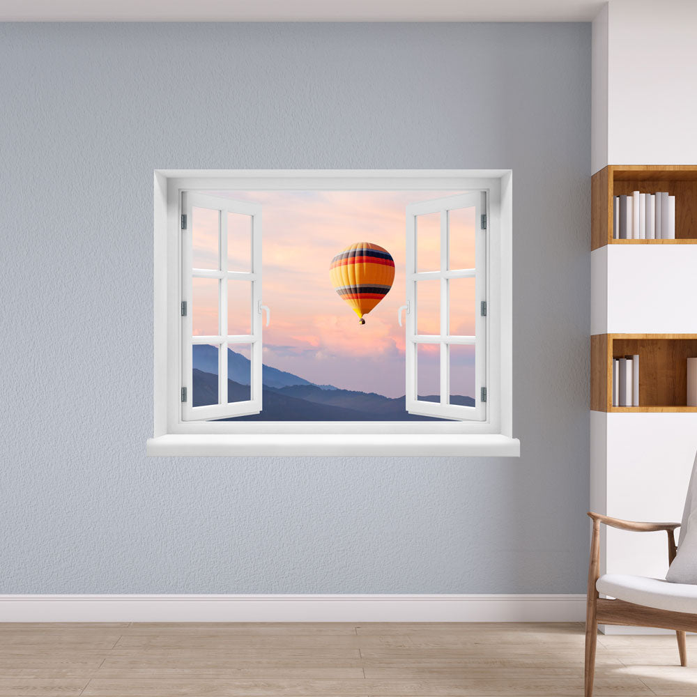 Wandaufkleber mit dem Motiv offenes Fenster mit Blick auf einen Heißluftballon im Sonnenuntergang über einer Berglandschaft – dekoratives 3D-Fensterbild mit Panorama-Effekt. Als Dekoidee an einer hellblauen Wand in einer Lesecke.
