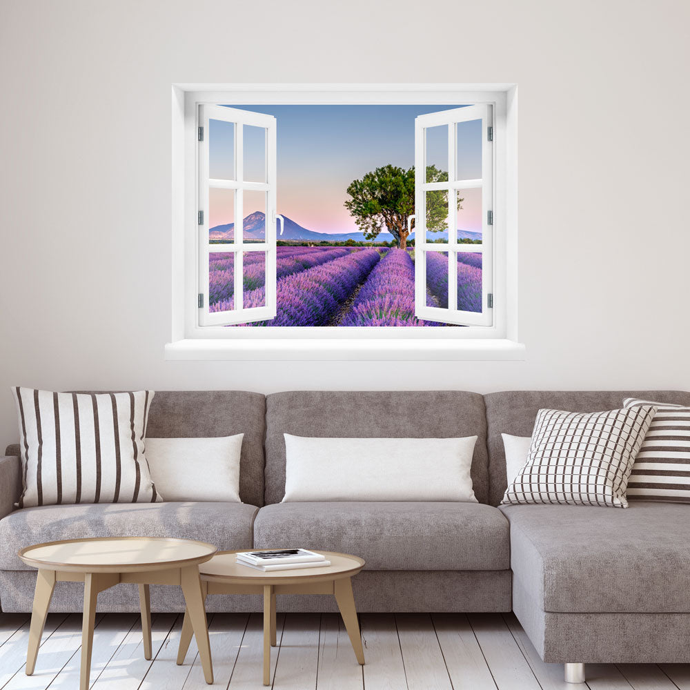 Wandaufkleber: offenes Fenster mit Blick auf ein weitläufiges Lavendelfeld bei Sonnenuntergang, mit einem markanten Baum in der Mitte und sanften Bergen im Hintergrund – romantische Landschaft in Pastellfarben. Als Dekoidee in einem Wohnzimmer, über einer grauen Couch an der der weissen Wand verklebt.
