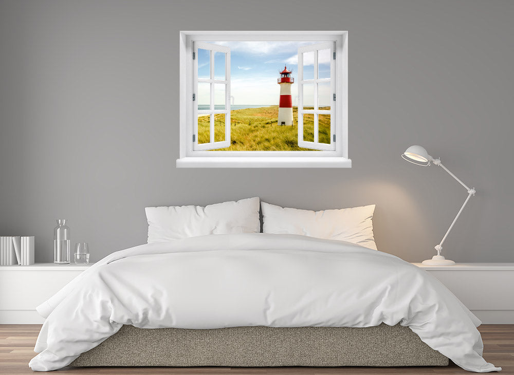 Großansicht eines Wandaufklebers: offenes Fenster mit Blick auf rot-weißen Leuchtturm inmitten von Dünenlandschaft an der Küste unter blauem Himmel – maritimes Küstenmotiv mit Nordsee- oder Ostsee-Flair. Als Einrichtungsbeispiel an einer grauen Wand, über einem Bett im Schlafzimmer verklebt.