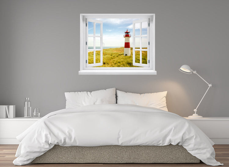Großansicht eines Wandaufklebers: offenes Fenster mit Blick auf rot-weißen Leuchtturm inmitten von Dünenlandschaft an der Küste unter blauem Himmel – maritimes Küstenmotiv mit Nordsee- oder Ostsee-Flair. Als Einrichtungsbeispiel an einer grauen Wand, über einem Bett im Schlafzimmer verklebt.