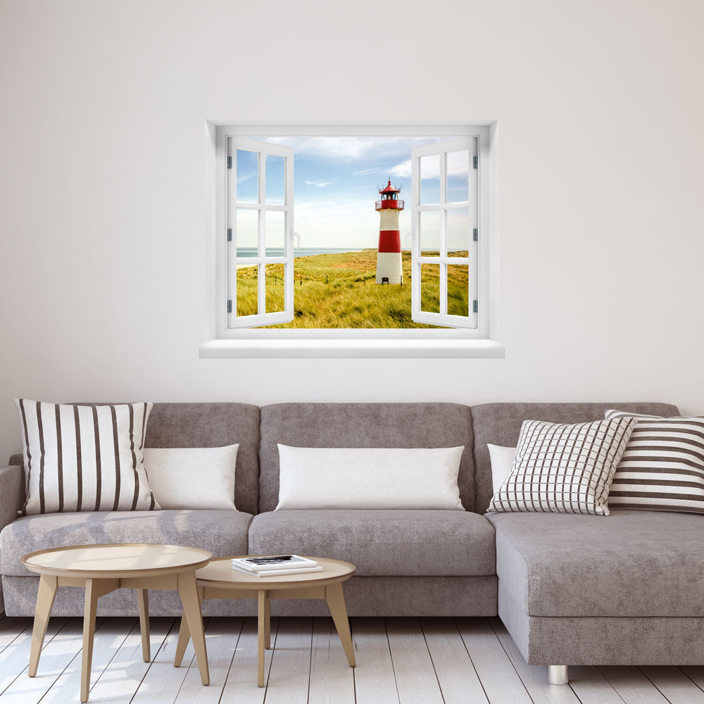 Großansicht eines Wandaufklebers: offenes Fenster mit Blick auf rot-weißen Leuchtturm inmitten von Dünenlandschaft an der Küste unter blauem Himmel – maritimes Küstenmotiv mit Nordsee- oder Ostsee-Flair. Als Einrichtungsbeispiel an einer weissen Wand, über grauen Couch im Wohnzimmer verklebt.