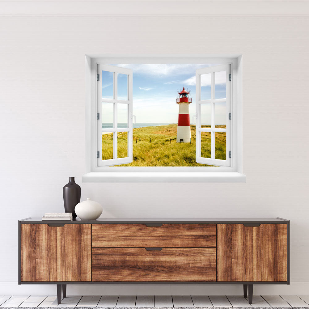 Großansicht eines Wandaufklebers: offenes Fenster mit Blick auf rot-weißen Leuchtturm inmitten von Dünenlandschaft an der Küste unter blauem Himmel – maritimes Küstenmotiv mit Nordsee- oder Ostsee-Flair. Als Einrichtungsbeispiel an einer weissen Wand, über einem Holz-Sideboard verklebt.
