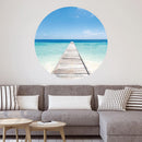 Großansicht eines runden Wandaufklebers mit einem Motiv vom Steg in der Karibik mit blauem Meer. Das Bild schafft einen 3D Effekt und sorgt für eine Tiefe. Als Dekoidee ist es an einer weissen Wand verklebt über einer grauen Couch.