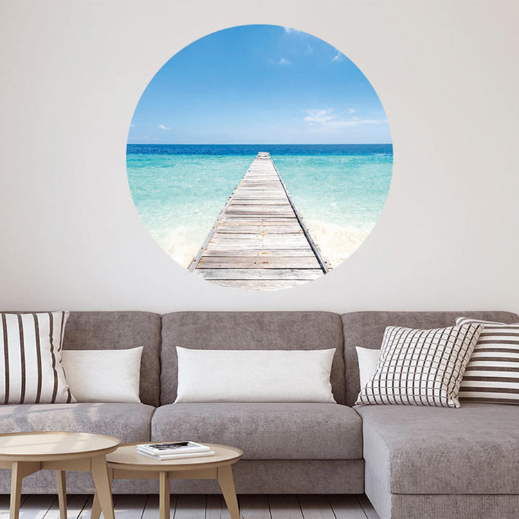 Großansicht eines runden Wandaufklebers mit einem Motiv vom Steg in der Karibik mit blauem Meer. Das Bild schafft einen 3D Effekt und sorgt für eine Tiefe. Als Dekoidee ist es an einer weissen Wand verklebt über einer grauen Couch.