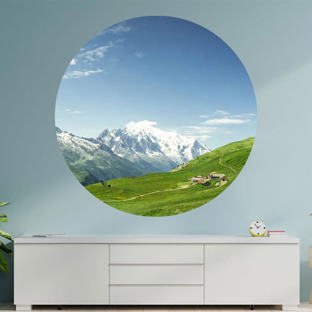 Ein großer, runder, Wandaufkleber mit einem Alpen-Wiesen-Motiv ist an einer Wand verklebt. Es ist eine Dekoidee über einer weissen Kommode und für verschieden Räume möglich, die einen 3D Effekt schaffen.