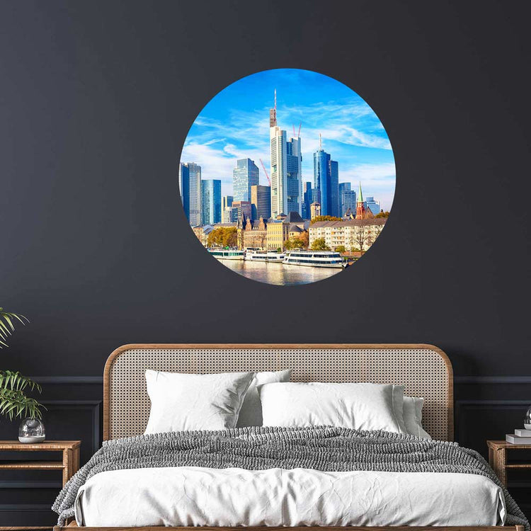 Rundes Wandbild mit der Skyline von Frankfurt am Main, stilvoll über einem Bett mit Rattan-Kopfteil auf dunkler Wand dekoriert.