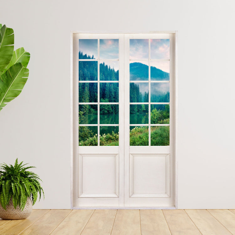 Dekorative Wohnraumgestaltung mit selbstklebendem Wandtattoo in der Optik einer Terrassentür mit dem Motiv: "See am Tannenwald", neben großen Zimmerpflanzen an einer Wand.