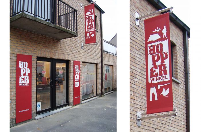 Zwei große Werbebanner hängen an der Fassade eines Gebäudes. Auf den Bannern steht "HOPPER WINKEL" in weißer Schrift, begleitet von stilisierten Grafiken, darunter eine Person und ein Zelt. Das Banner ist rot mit weißem Text und befindet sich an den Wänden des Geschäftsgebäudes.